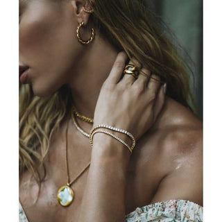Close-up of a woman's neck and arm wearing gold jewelry including a necklace, hoop earrings, and rings.