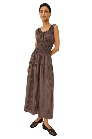 A woman modeling a sleeveless, smocked-bust midi dress with vertical pleats in a muted brownish-gray color, paired with black studded flats, against a plain white studio background.
