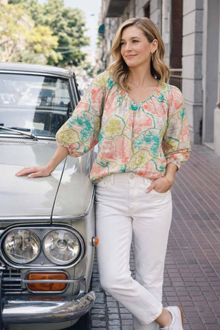 Woman in a floral blouse and white pants standing next to a vintage car on a city street.