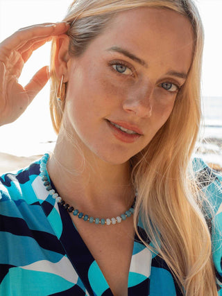 Woman with blonde hair and blue eyes wearing a colorful top and necklace, outdoors.
