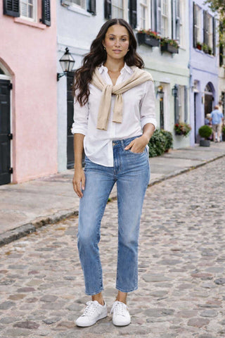 Woman wearing a white shirt, blue jeans, and a beige scarf on a street with colorful buildings.