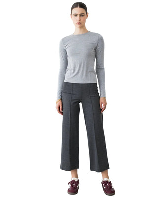 A model stands against a white wall wearing a light gray long-sleeve crewneck top, dark charcoal gray wide-leg cropped trousers, and burgundy lace-up shoes.