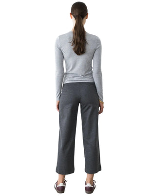 A woman viewed from behind wearing a light gray long-sleeve mock neck top and dark gray cropped wide-leg pants, standing against a plain white wall.