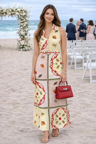 Woman in a patterned dress holding a red handbag on a beach with ocean and chairs in the background