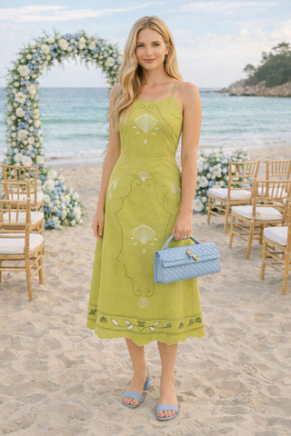 Woman in a green dress holding a blue handbag on a beach with floral decorations and chairs.