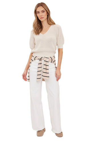 A woman modeling a light beige short-sleeve knit top, white wide-leg pants, and a brown and white striped tie belt, standing against a plain off-white studio background.