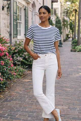 Woman wearing a striped shirt and white pants walking on a brick path with flowers and buildings in the background.