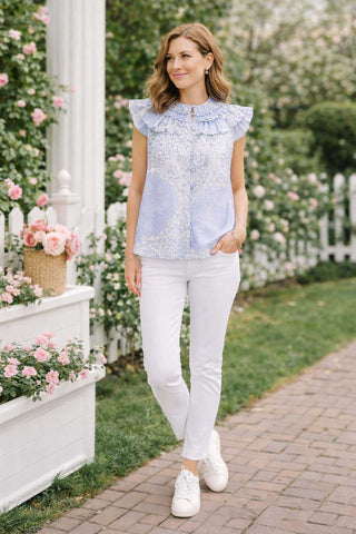 Woman in a light blue floral blouse and white pants walking outdoors with flowers and a white picket fence in the background.