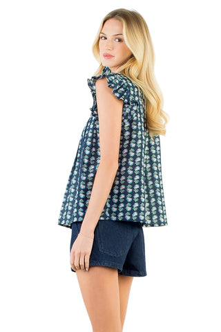 A blonde woman shown from the side/back wearing a sleeveless dark blue patterned top with ruffled shoulders and dark denim shorts, against a plain white studio background.