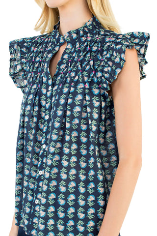 A close-up of a woman wearing a sleeveless blouse with a dark navy blue background, a dense small floral print in light blue, white, and green, and ruffled flutter sleeves with keyhole cutouts at the collar.