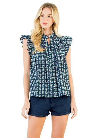 A blonde woman modeling a sleeveless button-front blouse with a ruffled tie-neck collar and an allover small floral print in blue and white, paired with dark denim shorts against a plain white studio background.