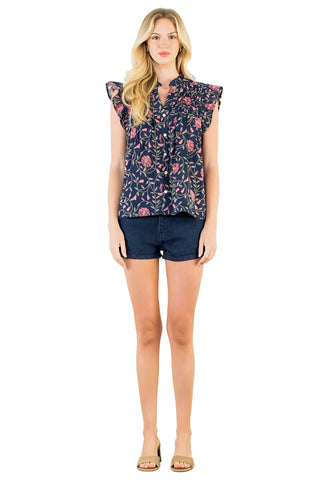 A woman modeling a sleeveless navy blue blouse with a pink and green floral print, paired with dark denim shorts, against a plain white background.