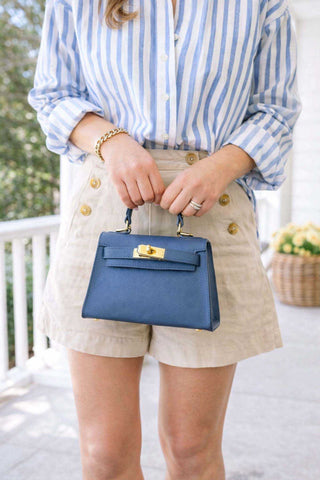 Person holding a blue handbag wearing a striped shirt and beige shorts on a blurred outdoor background