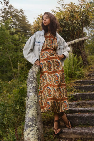 A woman stands on outdoor stone steps in a wooded setting, wearing a long-sleeve midi dress with an intricate orange, blue, and beige paisley pattern, styled with a light-wash denim jacket and brown strappy sandals.