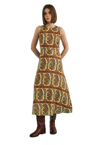 A woman modeling a sleeveless midi dress with a brown, beige, and multicolor paisley pattern, paired with brown leather boots, set against a solid white background.