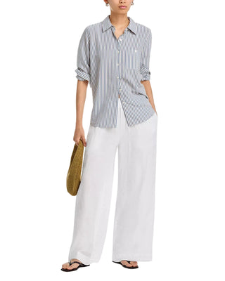 A woman models a light blue and white vertically striped button-down shirt with rolled sleeves, paired with wide-leg white trousers and holding a woven straw bag against a plain white background.
